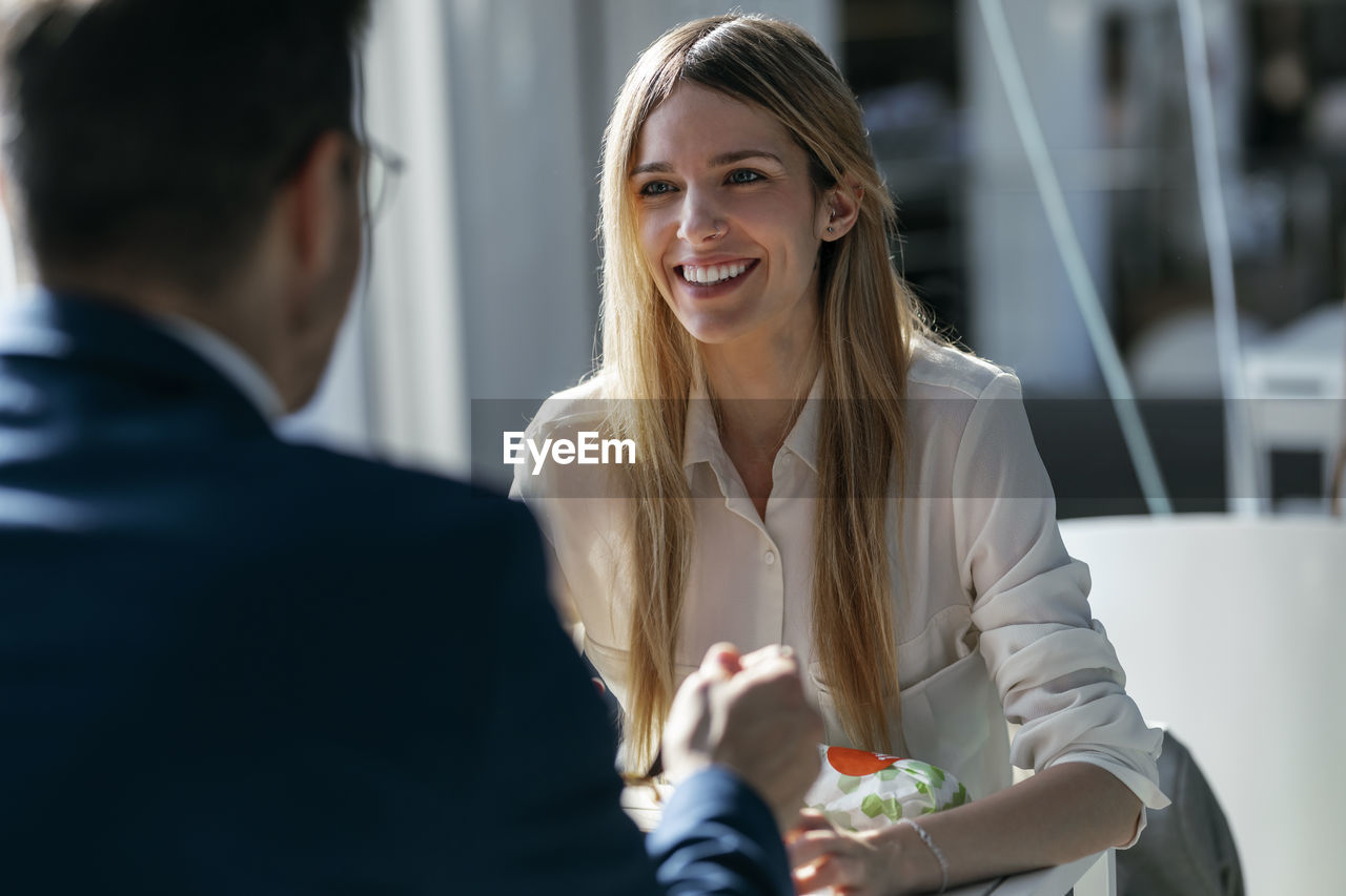 Smiling businesswoman having lunch with colleague while sitting in cafeteria at office