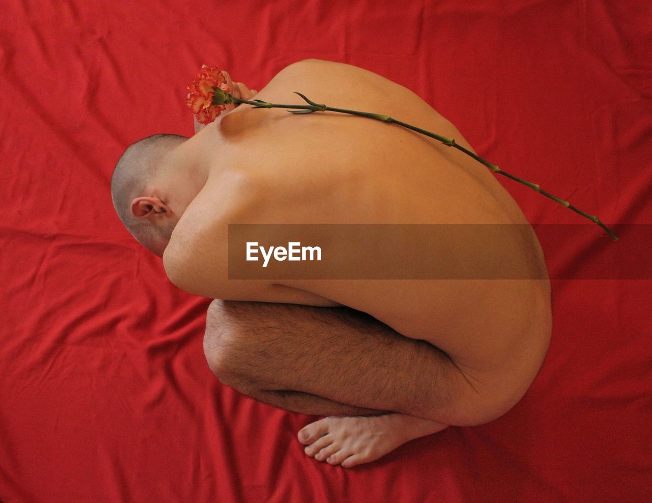 High angle view of naked man crouching with flower on red fabric