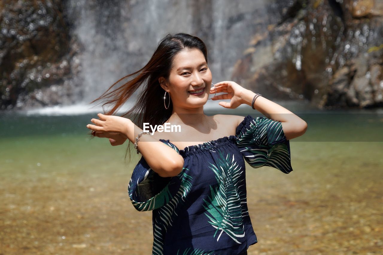 Portrait of smiling young woman standing against waterfall