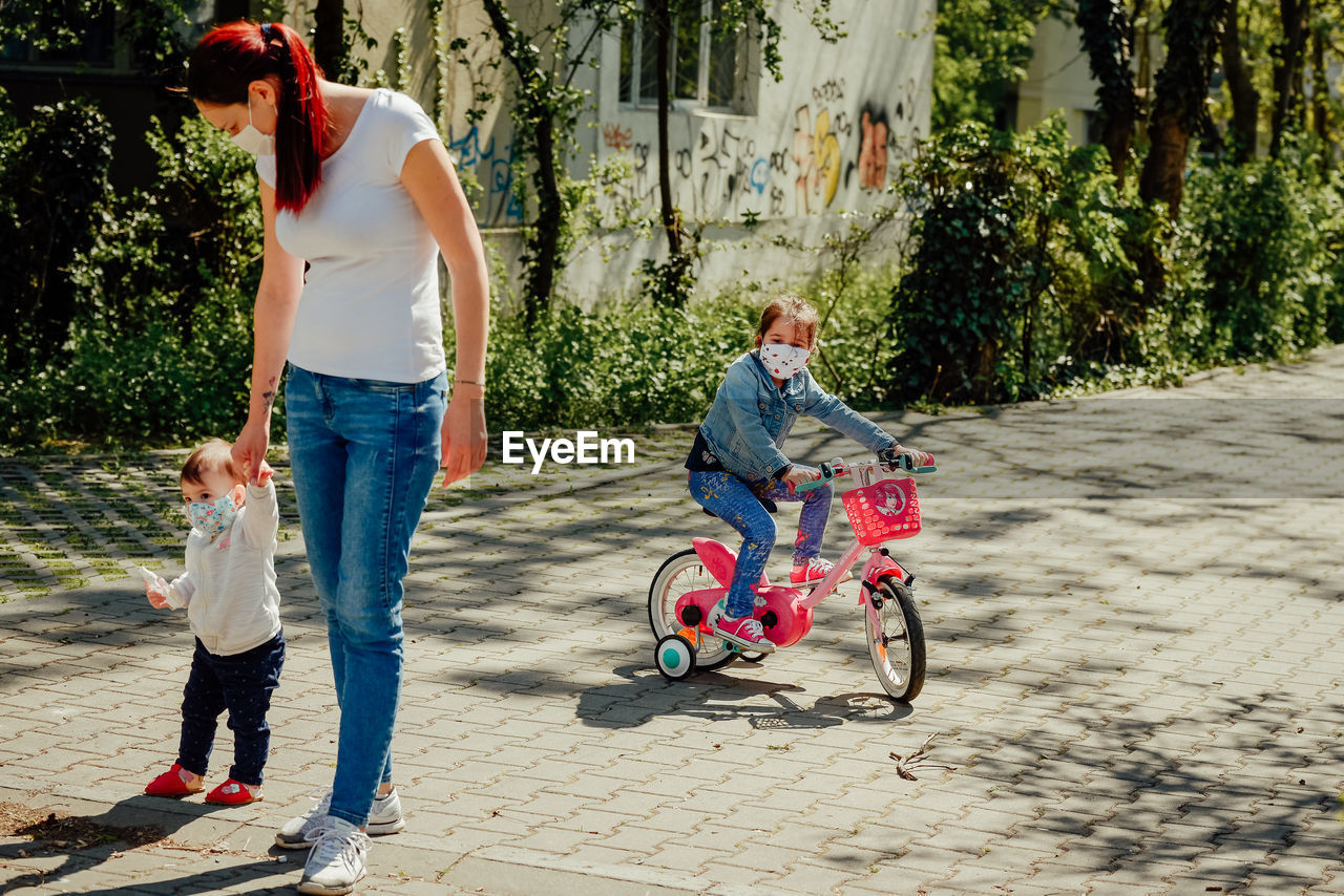Mother wearing a mask walking one daughter on foot and big sister on a bicycle pandemic year of 2021