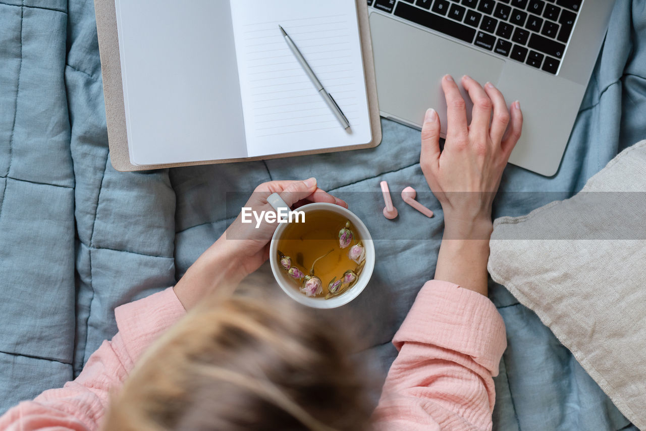 Overhead scene in pastel colors of a woman using a laptop, making notes, and drinking tea.