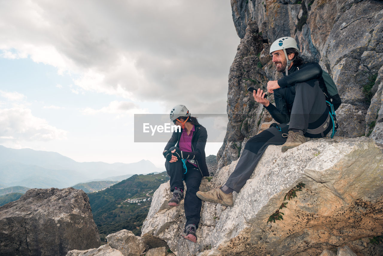 Concept: adventure. pair of climbers with helmet and harness. resting sitting on a rock. using the smart phone while looking at the horizon. via ferrata in the mountains.