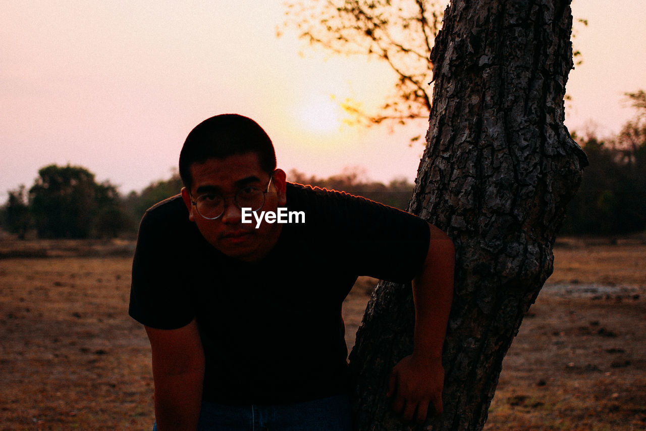 PORTRAIT OF MAN STANDING ON TREE TRUNK AGAINST SKY