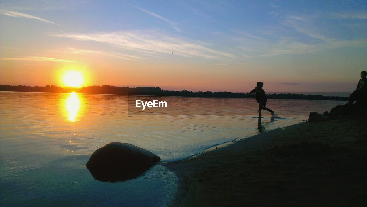 Silhouette boy playing on shore against sky during sunset