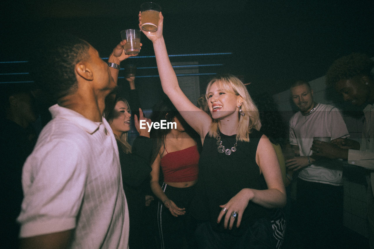 Happy young woman and man toasting drinks while dancing against friends at nightclub