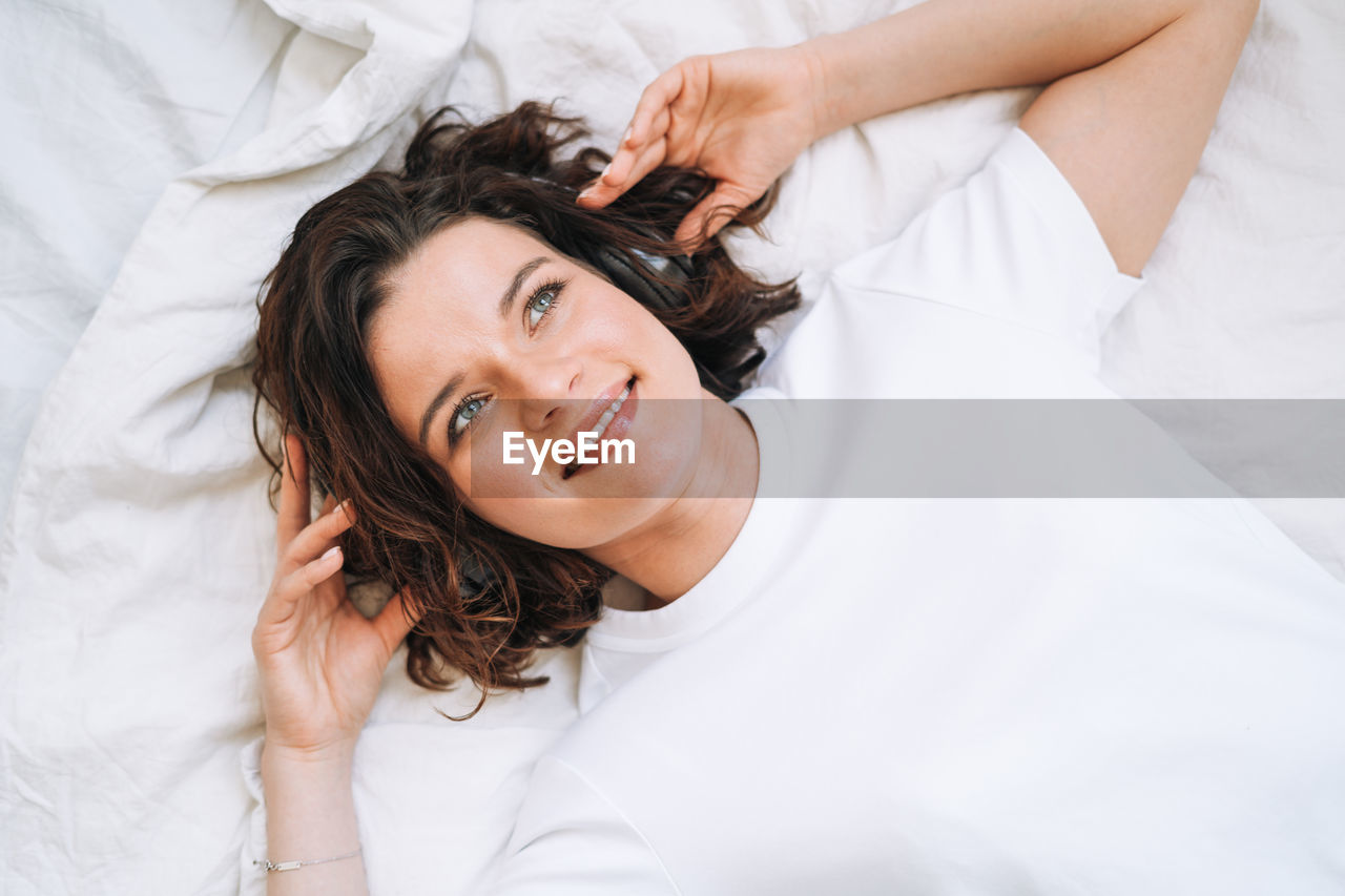 Young smiling brunette woman in headphones lying on bed at home, view from top