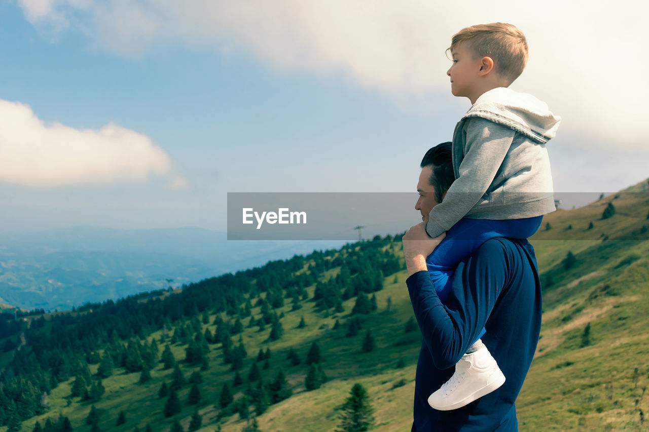 Happy father holding his son on the shoulders while enjoying at mountain peak. copy space.