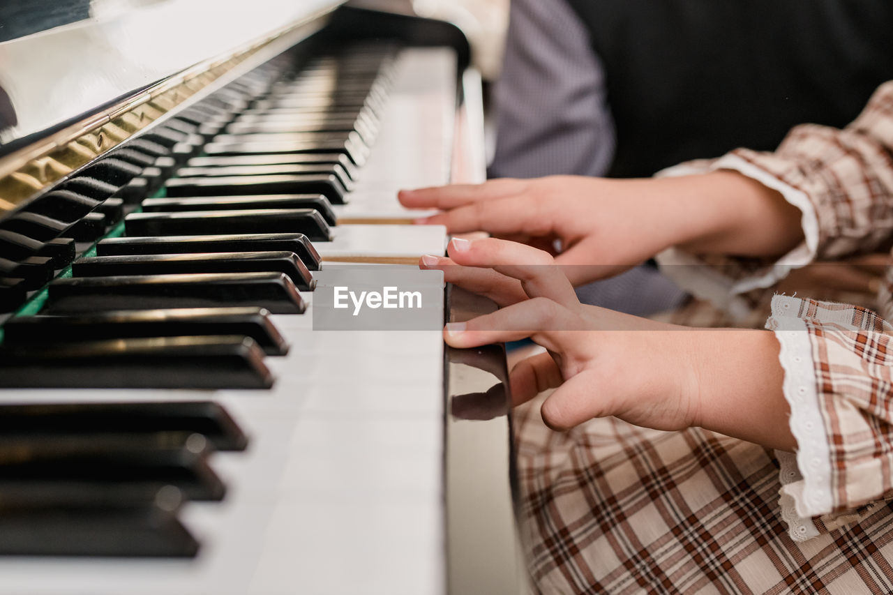 Crop unrecognizable child in checkered dress playing piano while having free time at home on blurred background