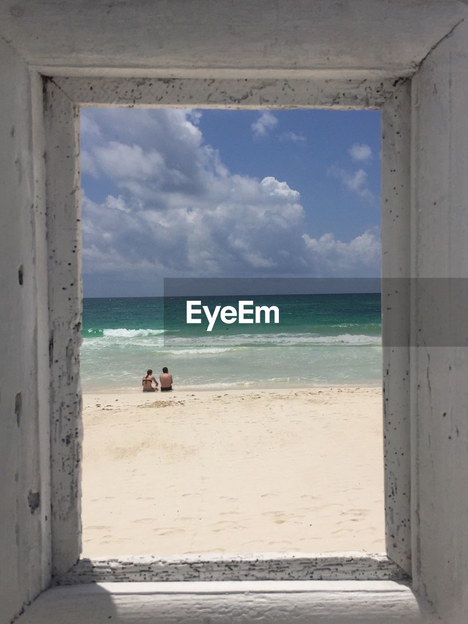 Couple sitting at beach seen through window against sky