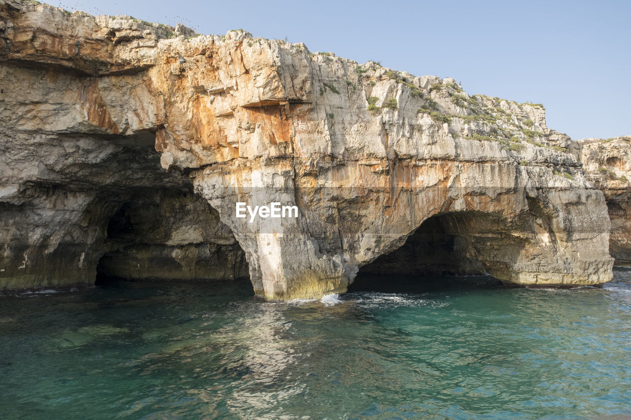 rock, water, cliff, cave, rock formation, sea cave, natural arch, coast, nature, sea, land, beauty in nature, formation, terrain, sky, scenics - nature, arch, no people, clear sky, geology, travel destinations, eroded, tranquility, non-urban scene, bay, beach, environment, travel, blue, wadi, outdoors, waterfront, day, idyllic, landscape, physical geography, tranquil scene, sunny, limestone, ocean, coastline, sunlight, tourism