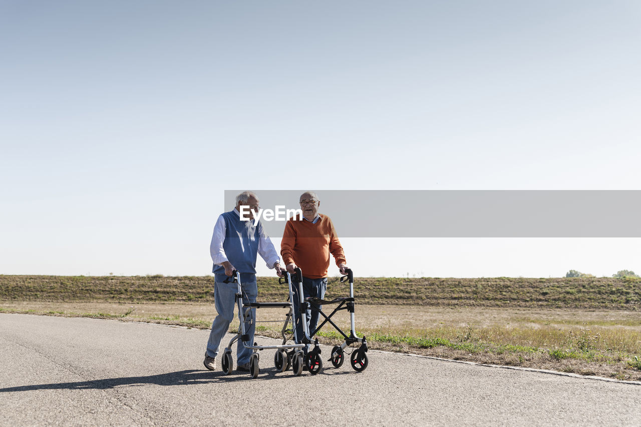 Two old friends walking on a country road, using wheeled walkers