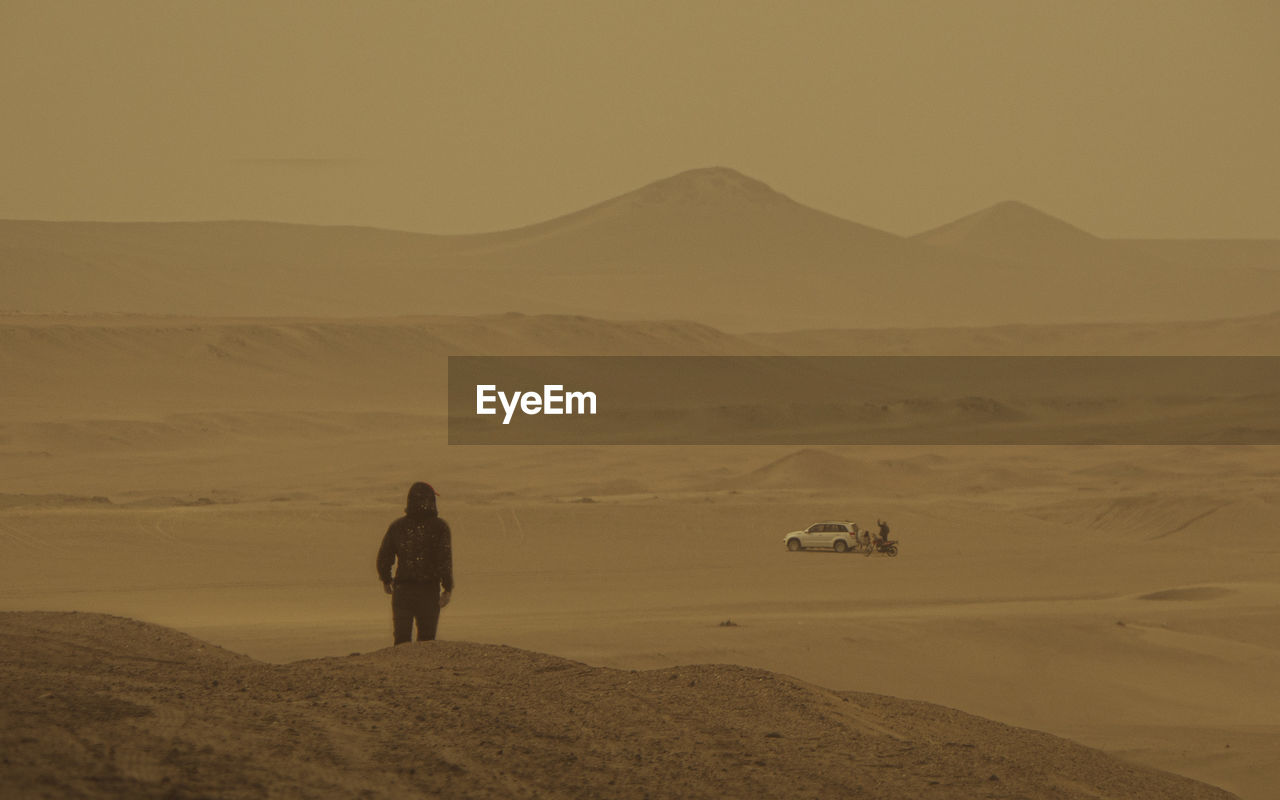 A lonely guy in the desert looks at a car in the distance. full length of man on desert against sky. 