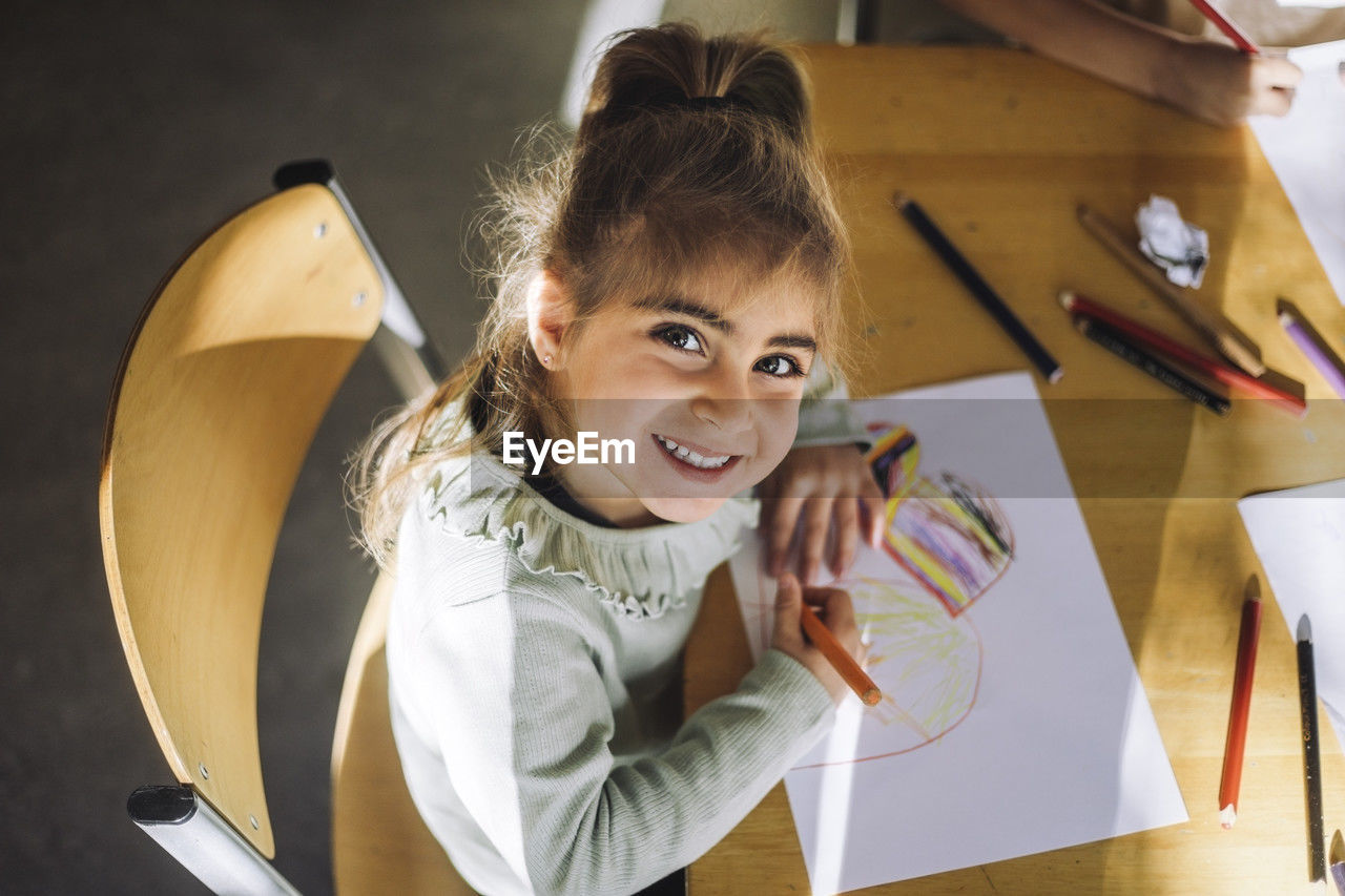 High angle view of smiling girl drawing with crayon on paper while sitting at bench in classroom