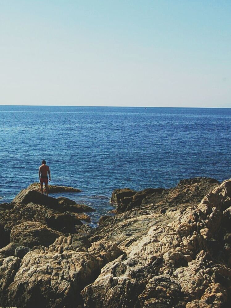 Rear view of man on rocks at sea against clear sky