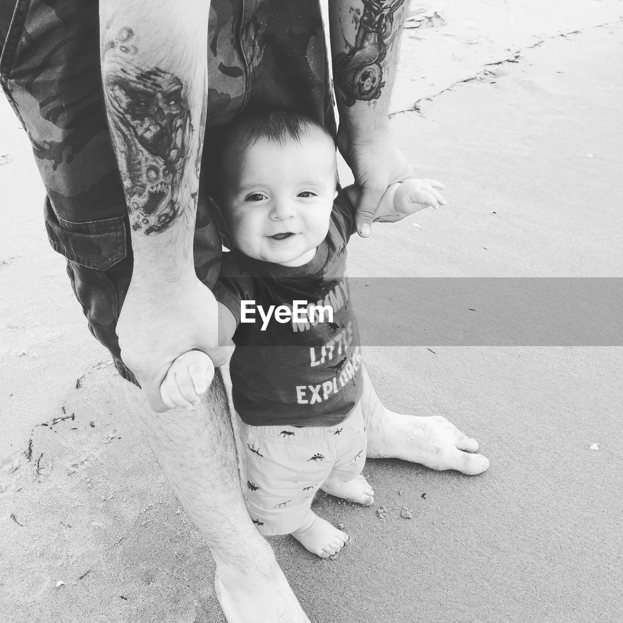 Low section of father holding cute baby boy while standing on sand at beach