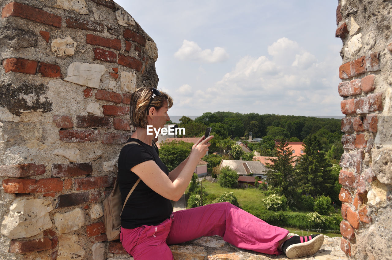 SIDE VIEW OF YOUNG WOMAN SITTING ON WALL AGAINST STONE WALLS