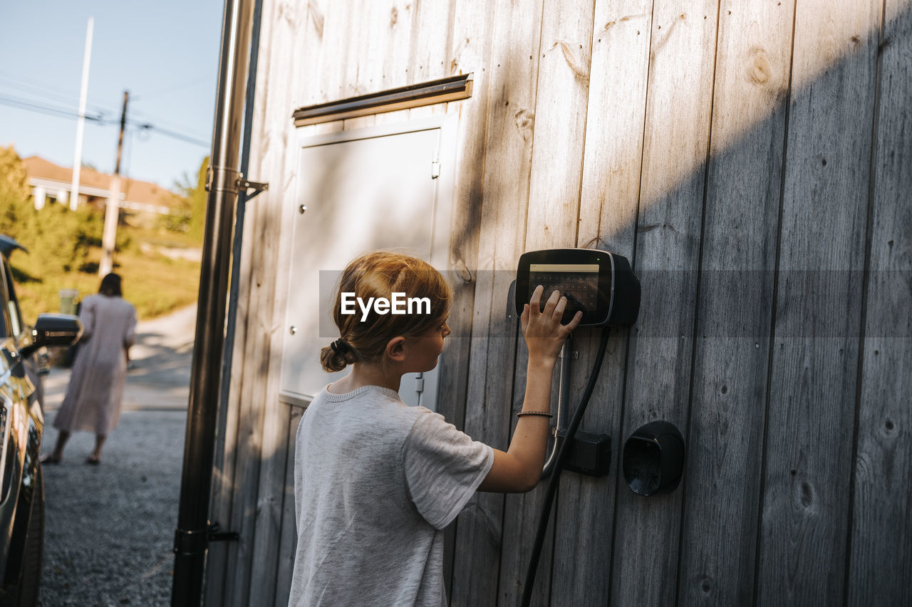 Boy with blond hair operating touch screen car charging device mounted on wooden wall