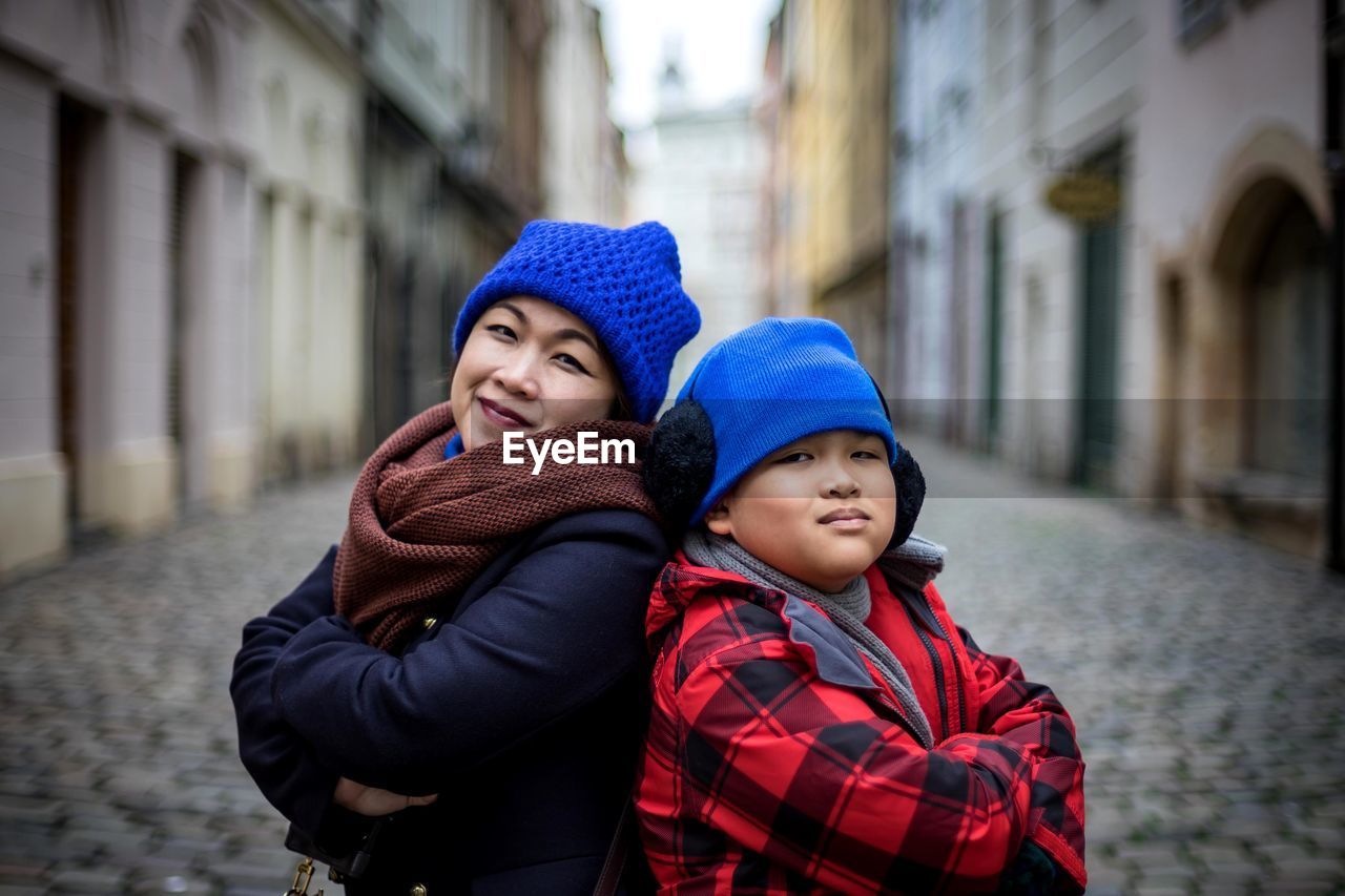 Portrait of smiling mother and son standing with arms crossed amidst buildings at city