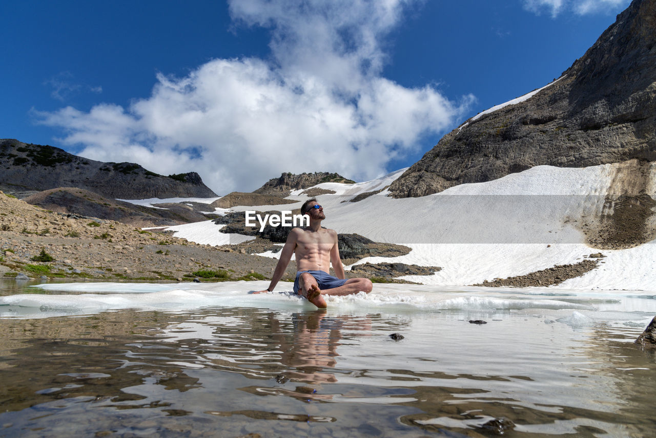 Full length of shirtless man sitting at lakeshore against sky