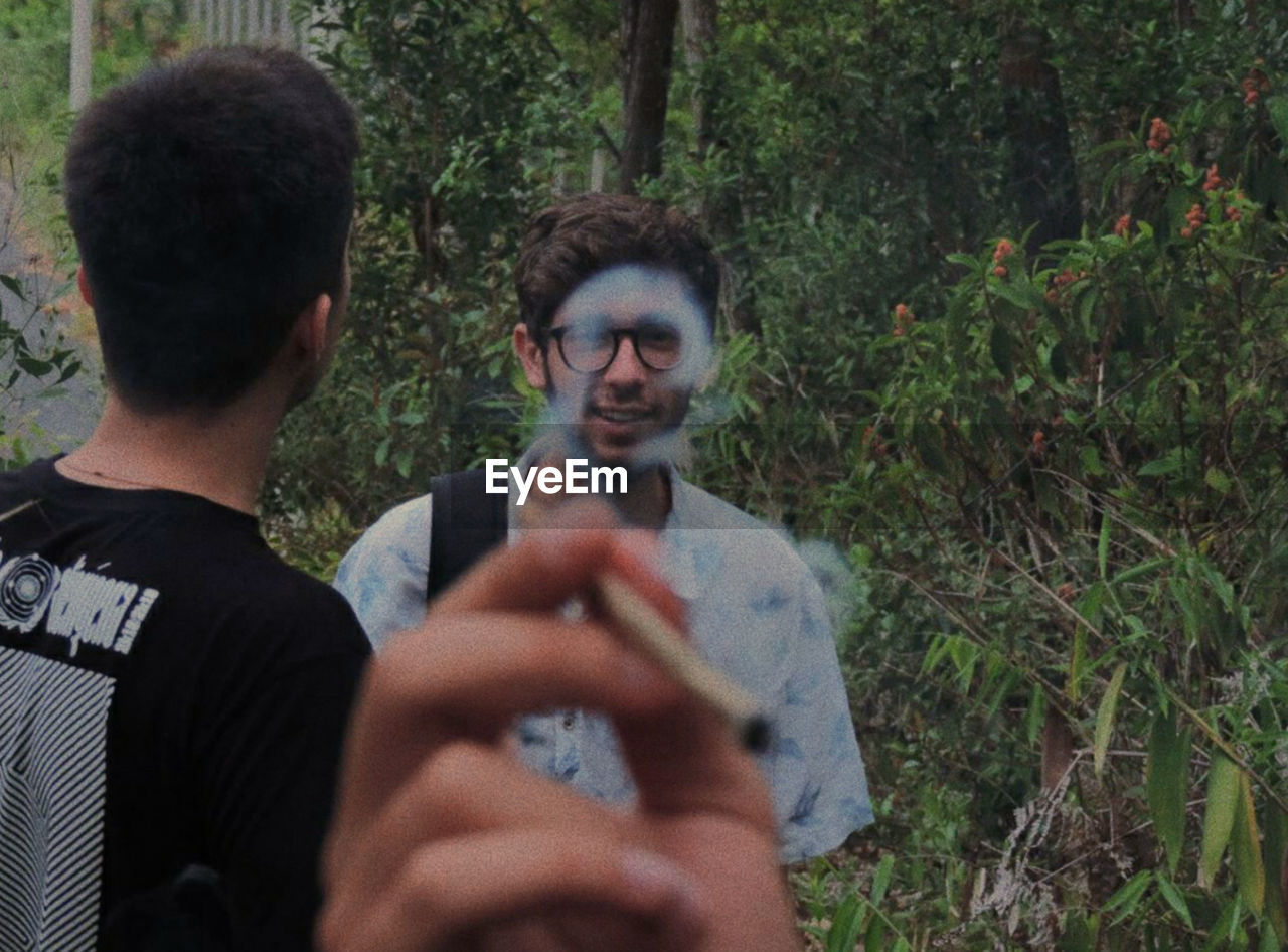 Cropped hand of man smoking cigarette with male friends standing against plants