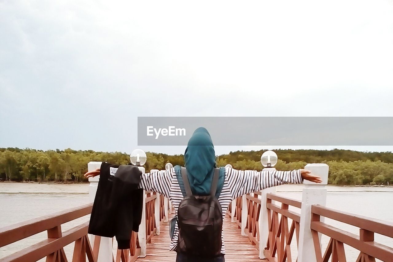 Rear view of woman with arms outstretched standing on pier over lake