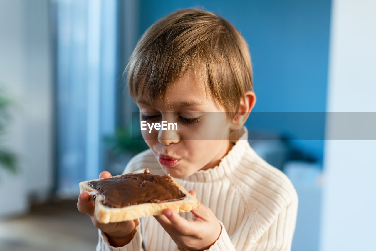 A boy bites into a sandwich with hazelnuts and cocoa, sitting on a table in the living room.