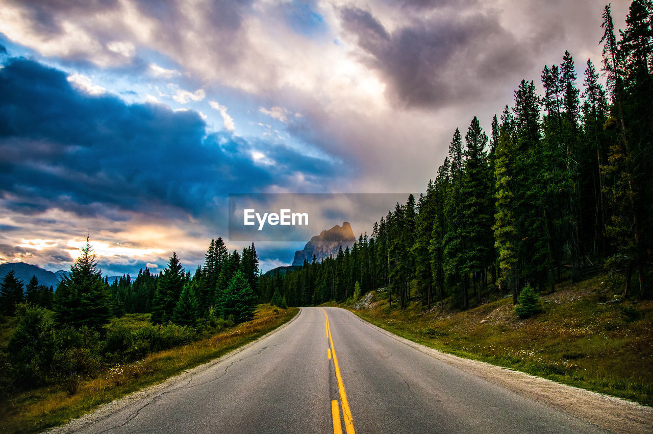 Empty road amidst trees against cloudy sky during sunset