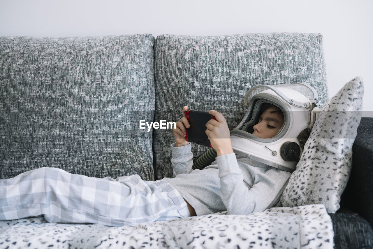 Adorable kid wearing pajama while sitting on couch. he is lying on couch while playing with a video game console at home