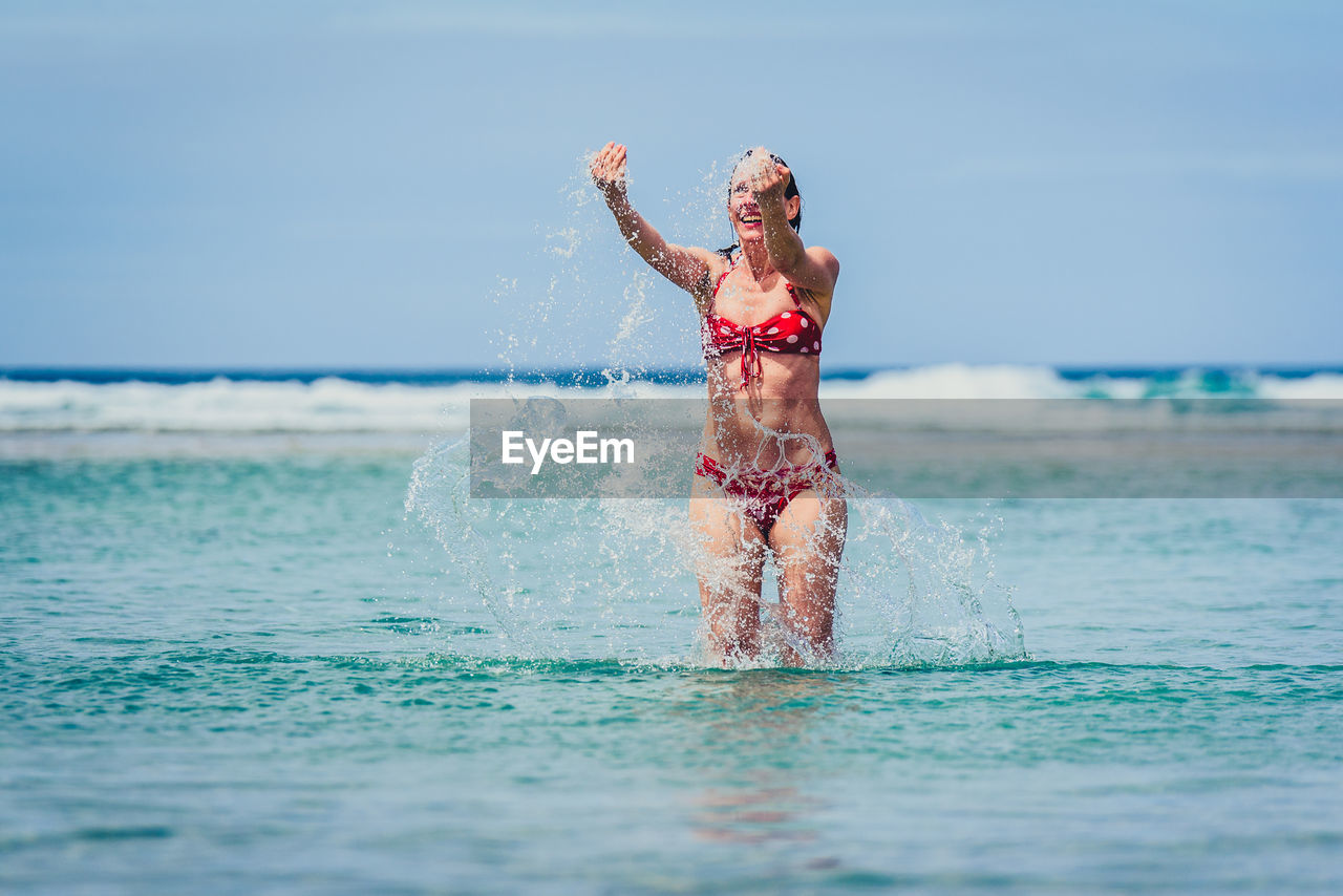 Happy mid adult woman splashing water in sea against sky