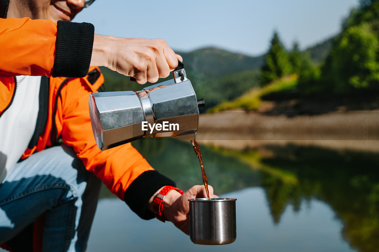 Midsection of man with coffee kettle pouring drink in cup standing by lake