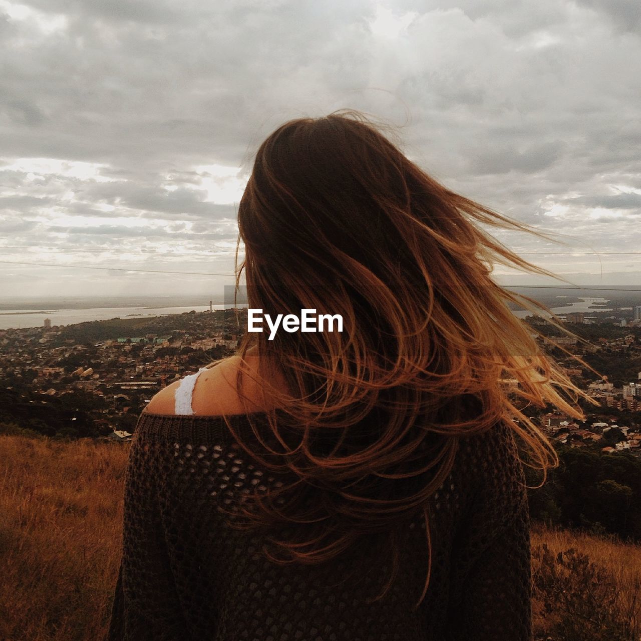 Rear view of woman with brown hair looking at cityscape while standing on field