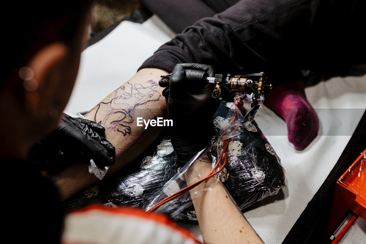 From above cropped unrecognizable man with using tattoo machine to make tattoo on leg of crop customer during work in salon