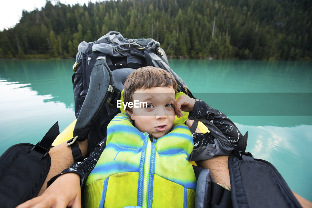 Boy wearing pfd rests against backpack on inflatable boat on blue lake