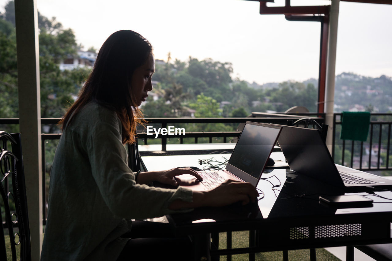 Side view of young female freelancer on vacation in casual clothing working with laptop at outdoors terrace of restaurant at sri lanka