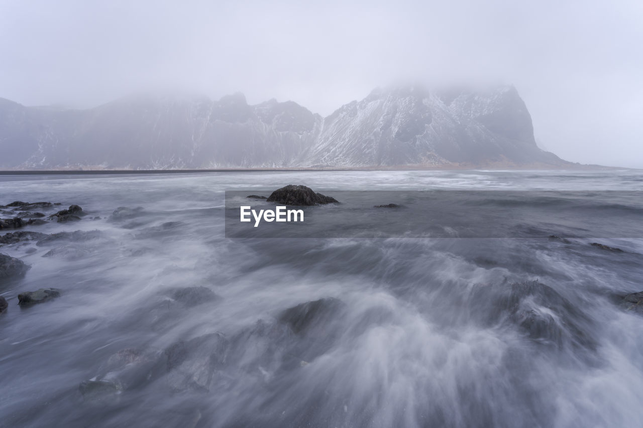 Picturesque scenery of powerful stormy sea water near rough rocky vestrahorn mountain formations on outcast misty day in stockness beach, iceland