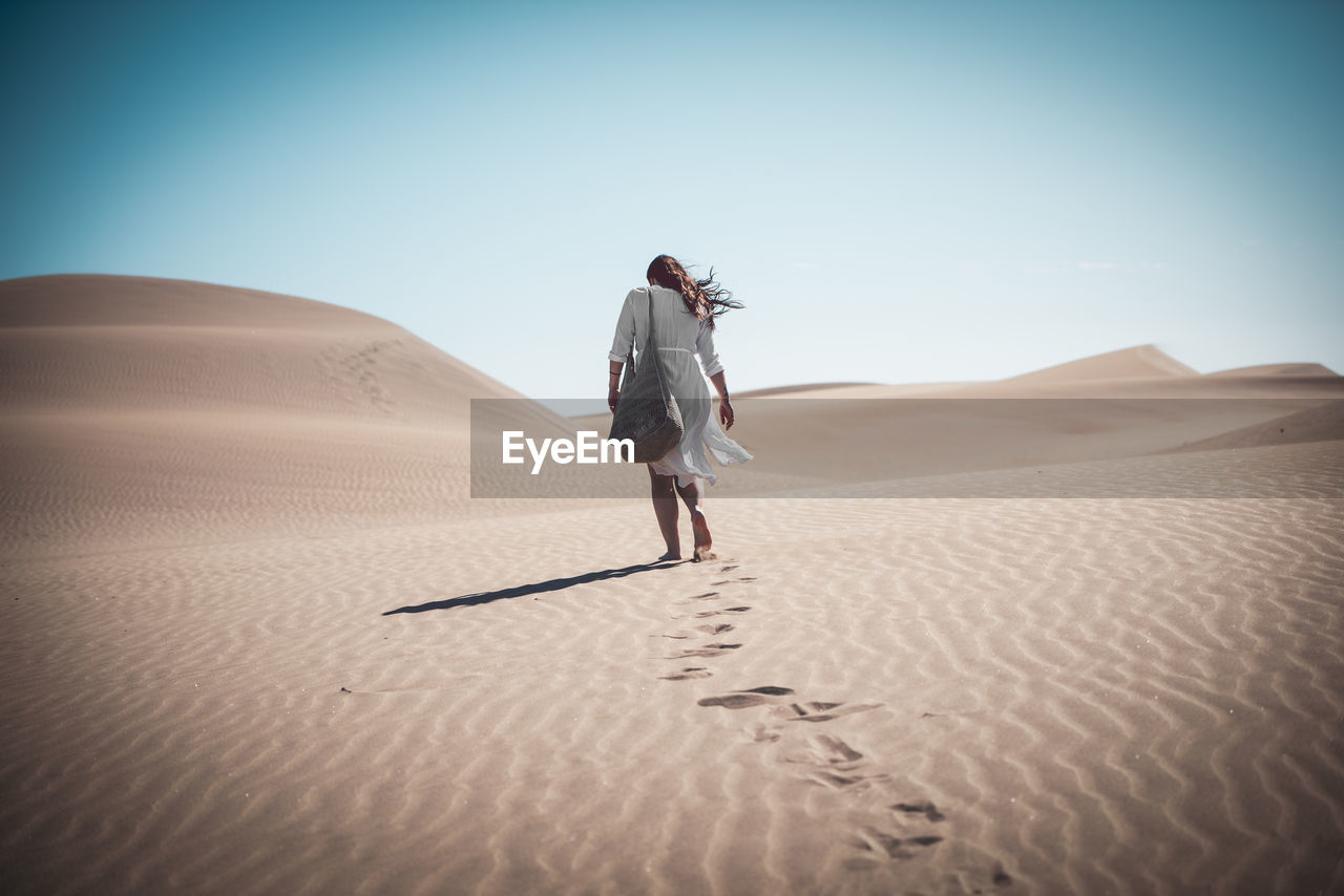 Rear view of woman walking on sand dune at desert against clear sky during sunny day