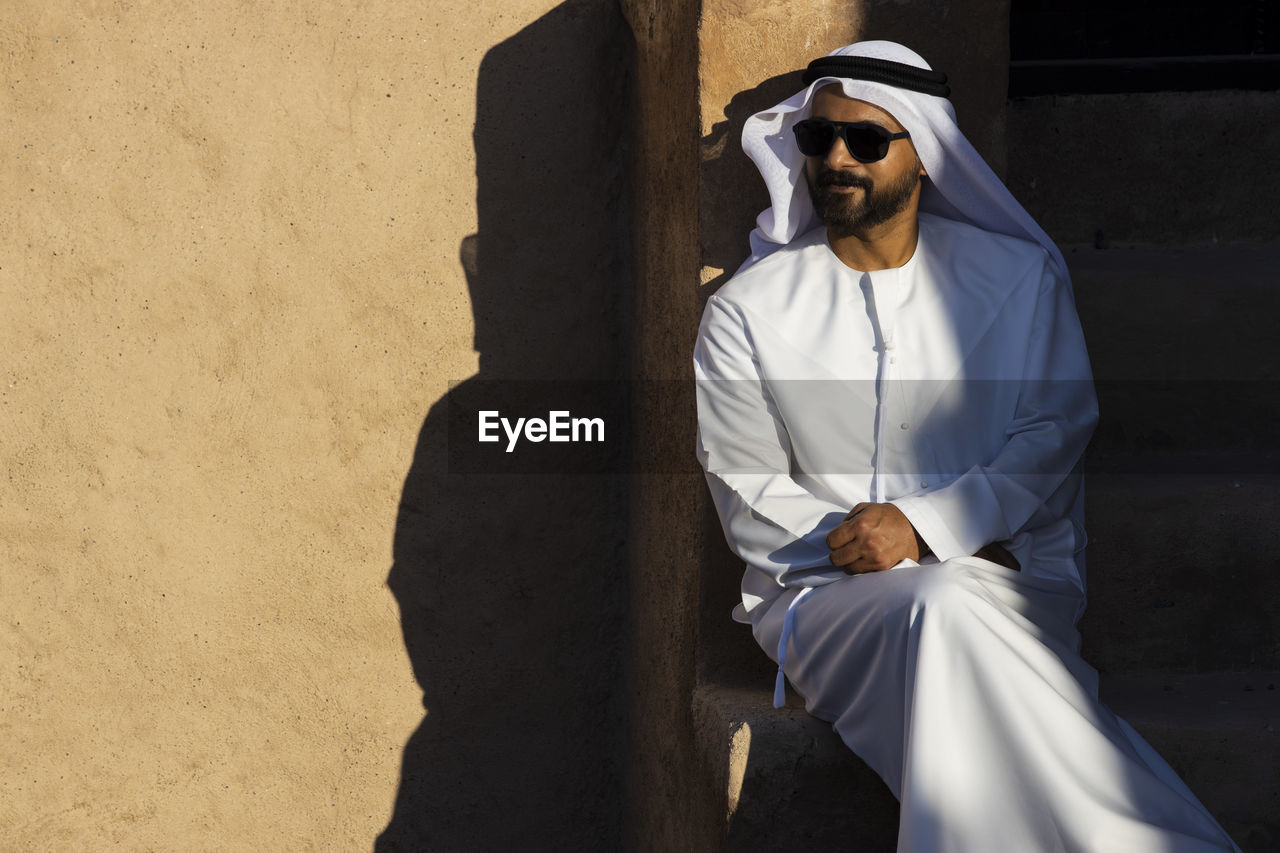 Man wearing traditional clothing and sunglasses sitting against wall