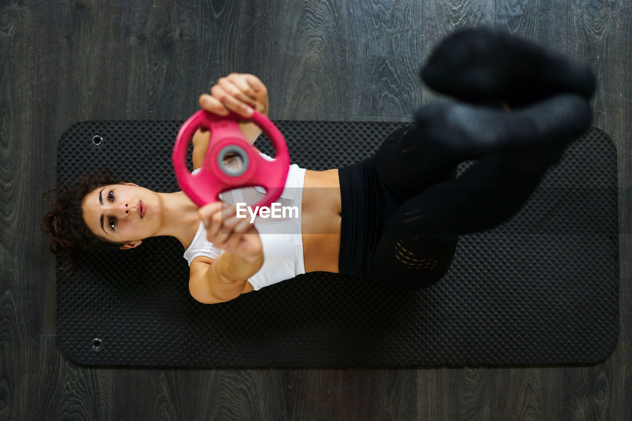 High angle view of young woman lifting weights while lying on mat in gym