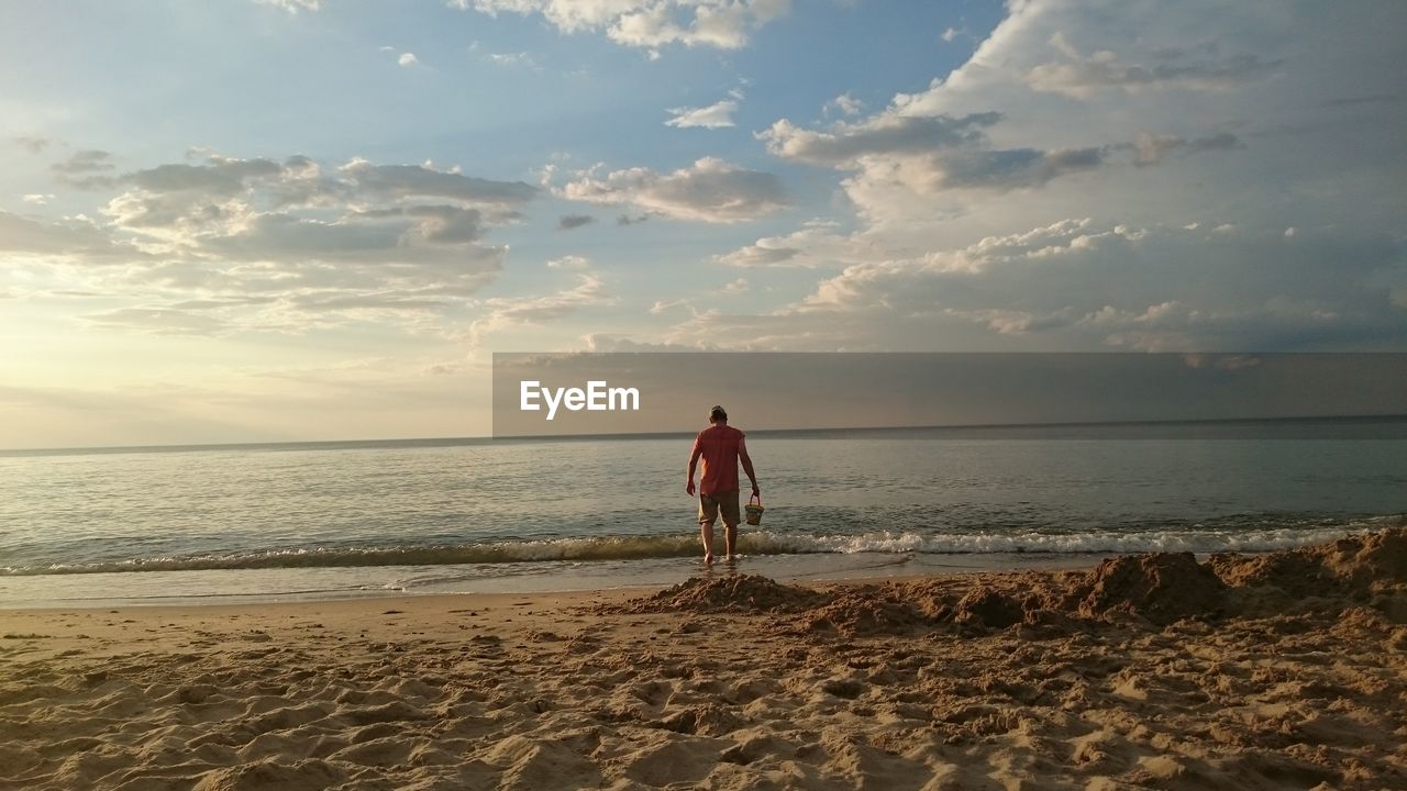 Rear view of man walking at beach against sky during sunset