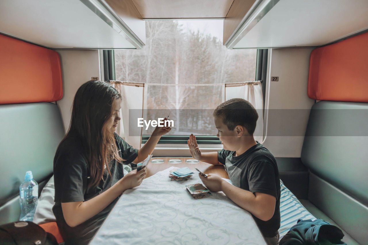 Sibling bonding, in train compartment brother and sister play card game, winter landscape in window