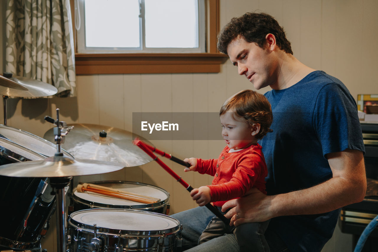 Father holding baby son playing drum at home