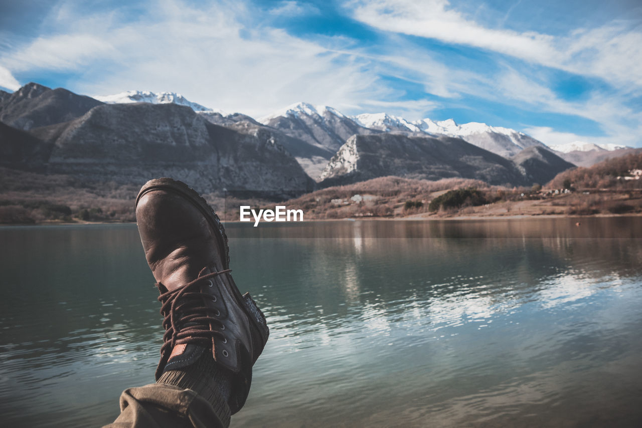 MAN STANDING BY LAKE AGAINST MOUNTAINS