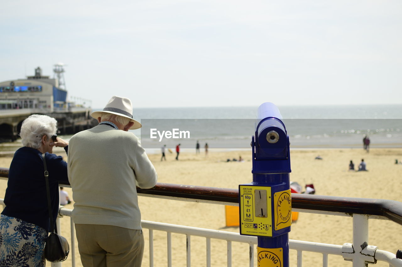 Senior couple standing by coin-operated binoculars at beach on sunny day