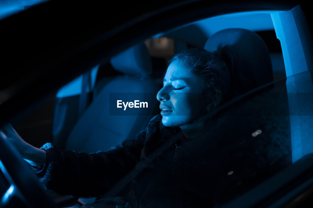 Side view of unhappy young female thinking about problems and crying while driving automobile at night