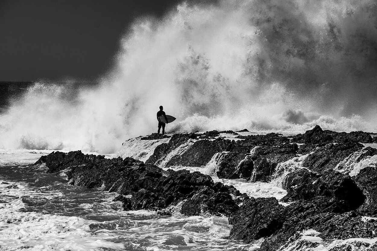 Silhouette man holding surfboard while standing against waves at rocky shore