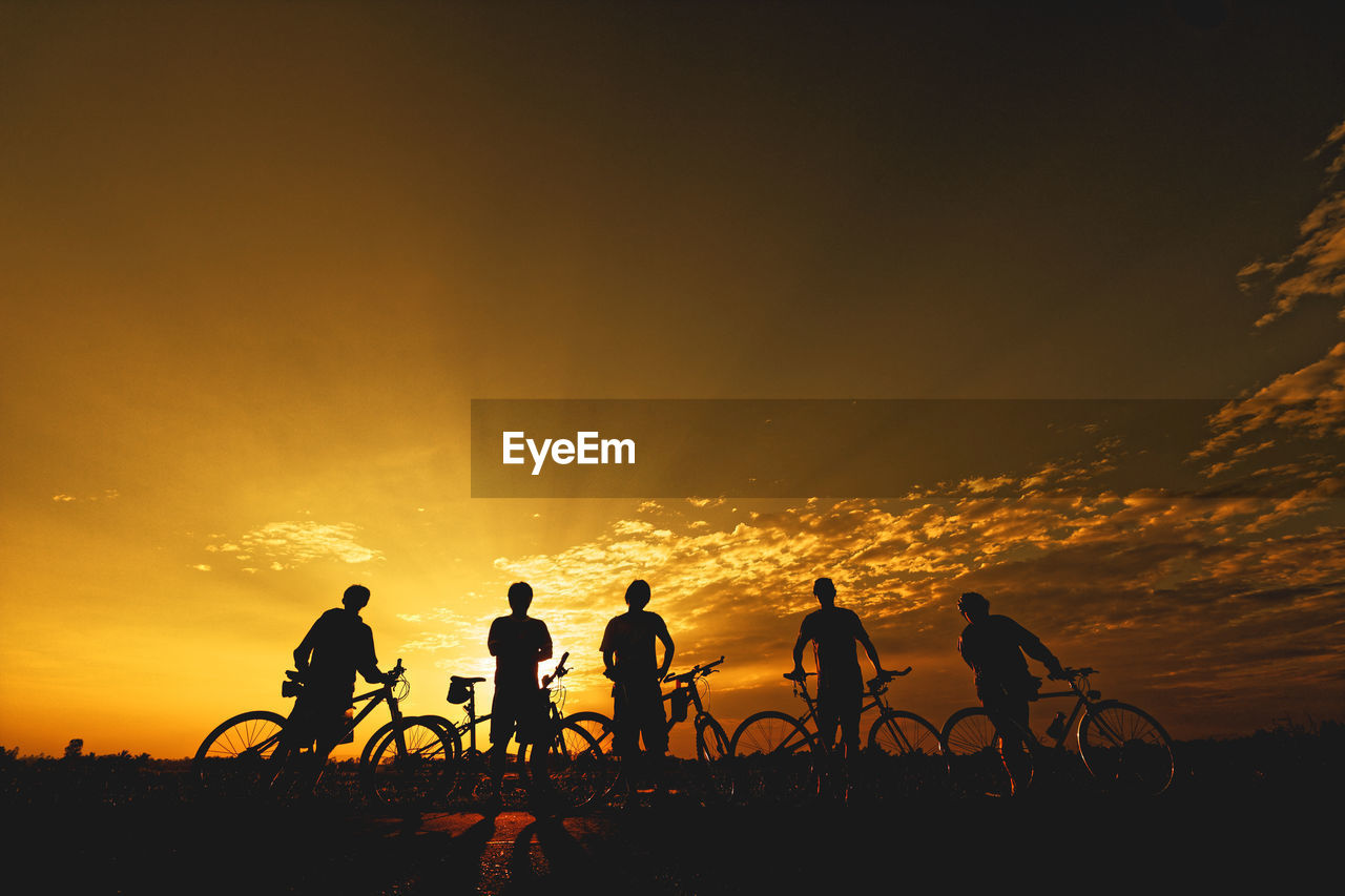 Silhouette people with bicycles standing on field against sky during sunset
