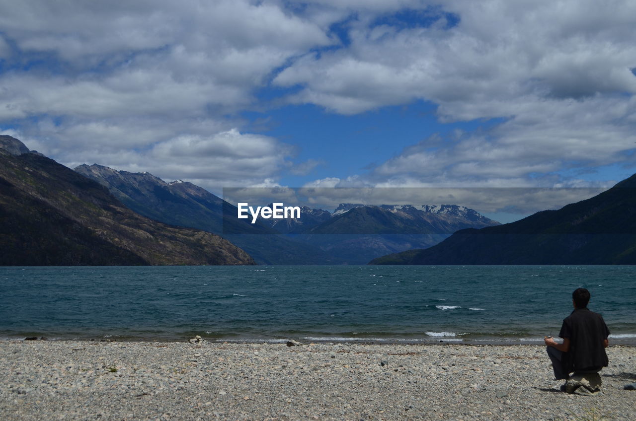 Man crouching at beach by mountains against cloudy sky