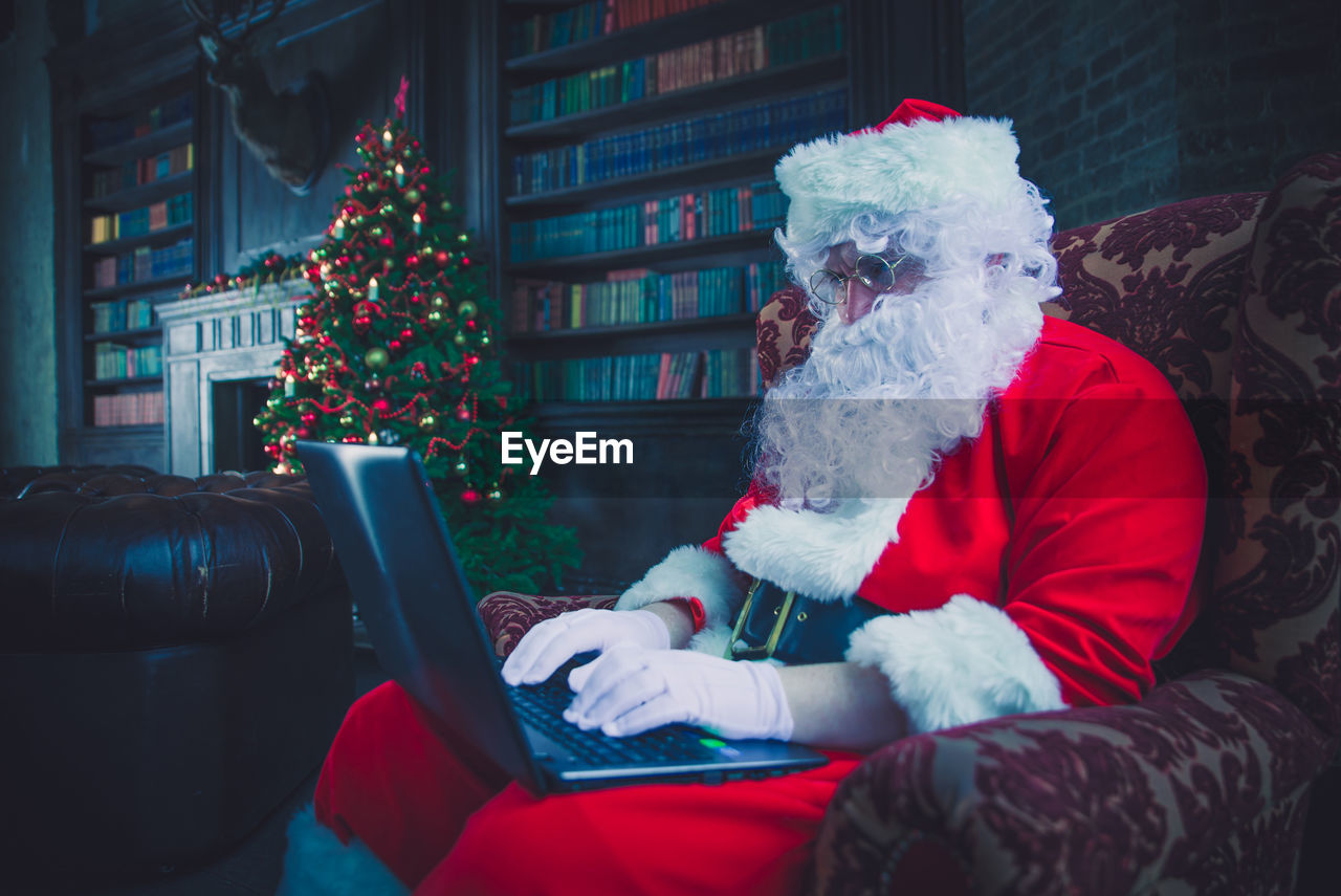 Man wearing santa claus costume using laptop on armchair at home