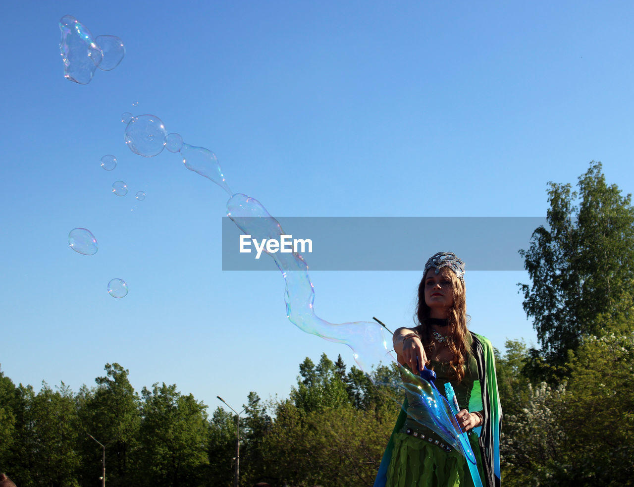 bubble, nature, blowing, one person, sky, bubble wand, blue, plant, women, fragility, toy, clear sky, mid-air, day, happiness, soap sud, childhood, tree, adult, fun, standing, sunny, motion, enjoyment, child, leisure activity, outdoors, flying, waist up, copy space, casual clothing, emotion, holding, carefree, sunlight, smiling, female, liquid bubble, young adult, looking, long hair, lifestyles, wind, low angle view, multi colored, hairstyle, environment, front view, summer, three quarter length, land