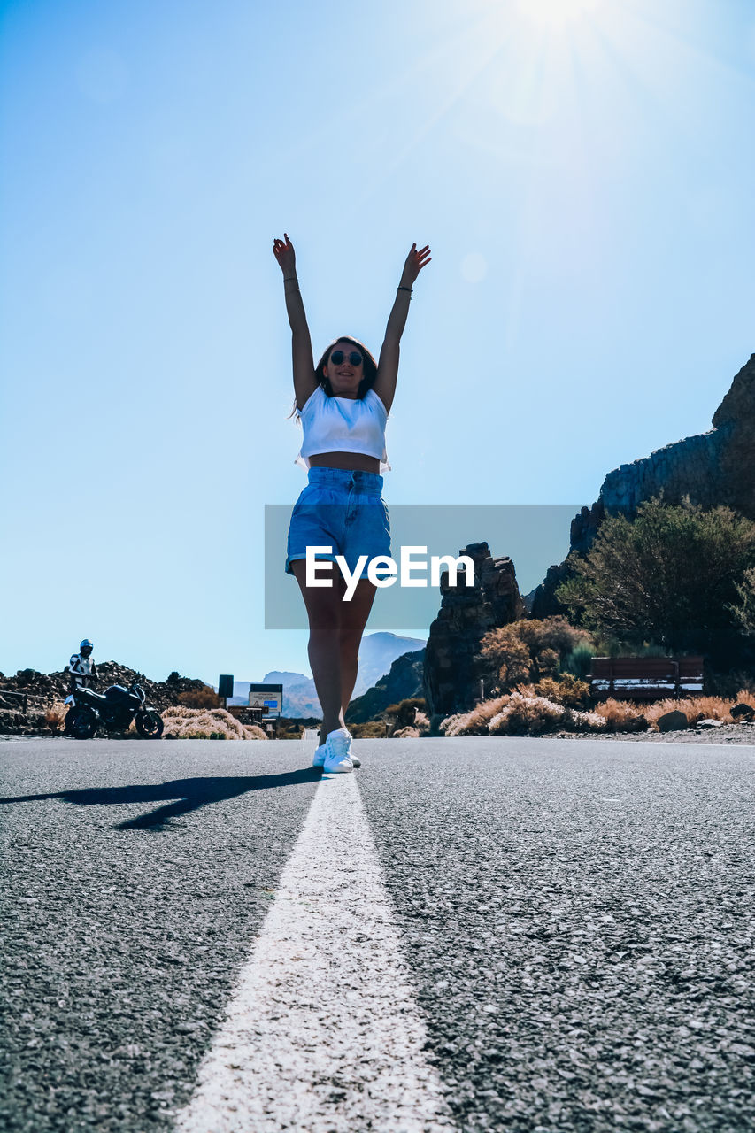 Full length of young woman standing on road against blue sky